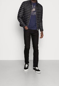 Chaqueta acolchada negra con cierre de cremallera, llevada sobre una camiseta de algodón azul marino, combinada con vaqueros negros y zapatillas negras con cordones blancos.