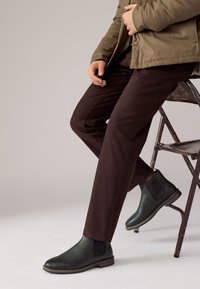 Brune bukser kombineret med sorte læderankelstøvler med elastiske sidepaneler og en tekstureret overflade, siddende på en metalstol.