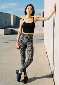 Tânără femeie în maiou negru și colanți cu imprimeu de leopard se sprijină de un perete de beton în aer liber, sub un cer senin și albastru.