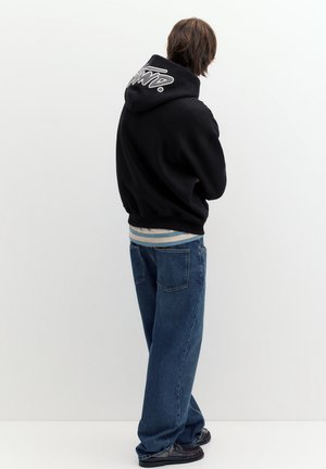 Person mit braunen Haaren, die einen schwarzen Hoodie mit weißem Text auf der Kapuze, blaue Jeans und schwarze Schuhe trägt und vor einem weißen Hintergrund steht.