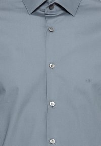 Camicia button-up grigia chiara realizzata in tessuto morbido. Presenta un colletto classico, sette bottoni e un logo ricamato discreto sul petto.