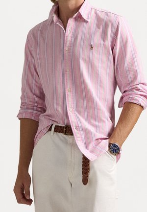 Man met een roze-wit gestreept overhemd met opgerolde mouwen, beige broek, gevlochten bruine riem en een blauw horloge.