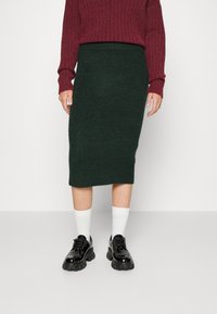 Jupe crayon en tricot vert foncé avec taille côtelée, associée à un pull côtelé bordeaux et des chaussures à lacets noires et épaisses. Chaussettes blanches visibles.
