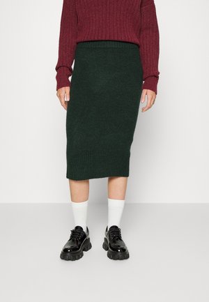 Dunkelgrüner Strickpencilrock mit geripptem Bund, kombiniert mit einem burgunderfarbenen, gerippten Pullover und schwarzen, klobigen Schnürschuhen. Weiße Socken sind sichtbar.