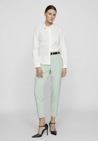 Chemise blanche à boutons, pantalon ajusté vert clair et chaussures à talons hauts noires. La tenue comprend une fine ceinture noire avec des accessoires en or.