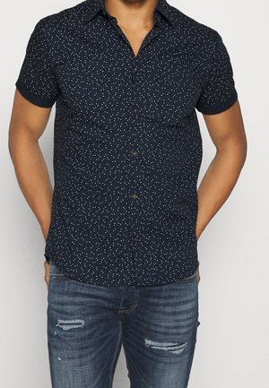 Man draagt een marineblauw overhemd met korte mouwen en witte spikkels, gecombineerd met beschadigde blauwe spijkerbroek, handen gedeeltelijk in de zakken.