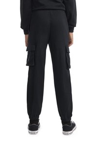 Μαύροι cargo παντελόνια από μαλακό ύφασμα, με πλευρικές τσέπες, ελαστικές μανσέτες και χαλαρή εφαρμογή, συνδυασμένα με μαύρα αθλητικά παπούτσια.