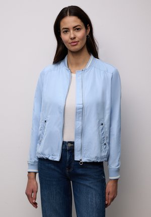 Donna con giacca bomber azzurra, camicia bianca e jeans blu, in piedi contro uno sfondo semplice con postura rilassata.
