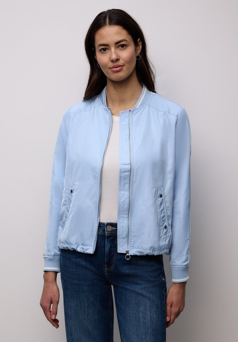 Mujer con chaqueta bomber azul claro, camisa blanca y vaqueros azules, de pie frente a un fondo liso con postura relajada.