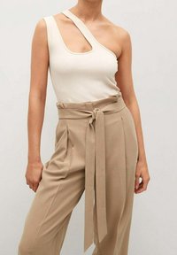 Kobieta ubrana w beżowe spodnie typu paperbag z wysokim stanem, wiązane paskiem, oraz asymetryczny top z kremowego żeberkowanego materiału na jedno ramię.