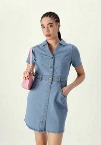 Γυναίκα με πλεξίδες, φορώντας ένα κοντομάνικο τζιν φόρεμα, κρατώντας μια μικρή ροζ τσάντα, με το ένα χέρι στην τσέπη, σε ουδέτερο φόντο.