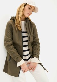 Olive green waterproof jacket with hood, zippers, and side pockets, layered over a cream and navy striped sweater. White cap visible.