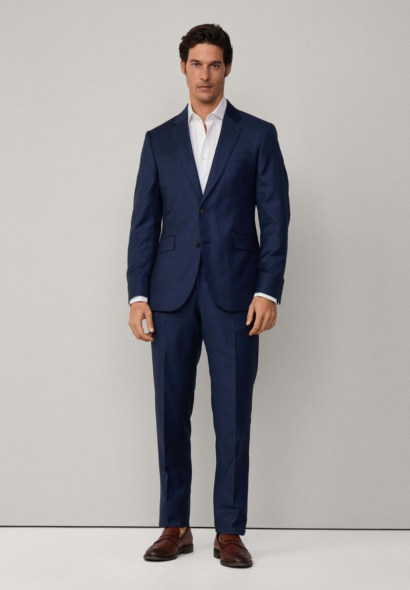 Costume bleu marine à la coupe ajustée, doté d'une fermeture à deux boutons, de revers crantés et de poches latérales. Assorti à une chemise blanche et des chaussures marron.