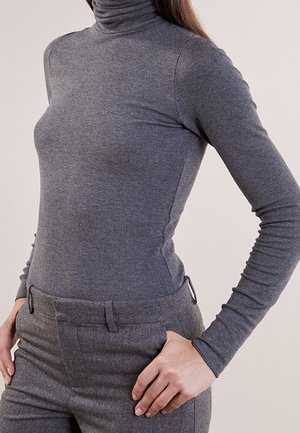 Person iført tætsiddende grå turtleneck sweater og grå højtaljede bukser med hænderne hvilende på hofterne foran en ensfarvet baggrund.