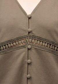 Κορυφή από ύφασμα τόουπ με V-λαιμόκοψη, που διαθέτει οριζόντια δαντελένια λωρίδα και κάθετες κουμπότρυπες καλυμμένες με ύφασμα στη μέση.