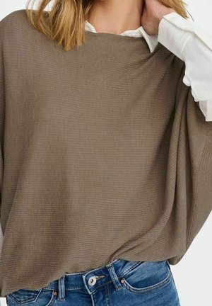 Vrouw die een taupekleurige geribbelde poncho draagt over een wit shirt en blauwe spijkerbroek, met één hand die haar nek aanraakt.