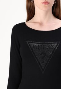 Juoda ilgo rankovės megztinis su deimanto formos, blizgučiais papuoštu logotipu, kuriame užrašyta "GUESS" ir klaustukas.