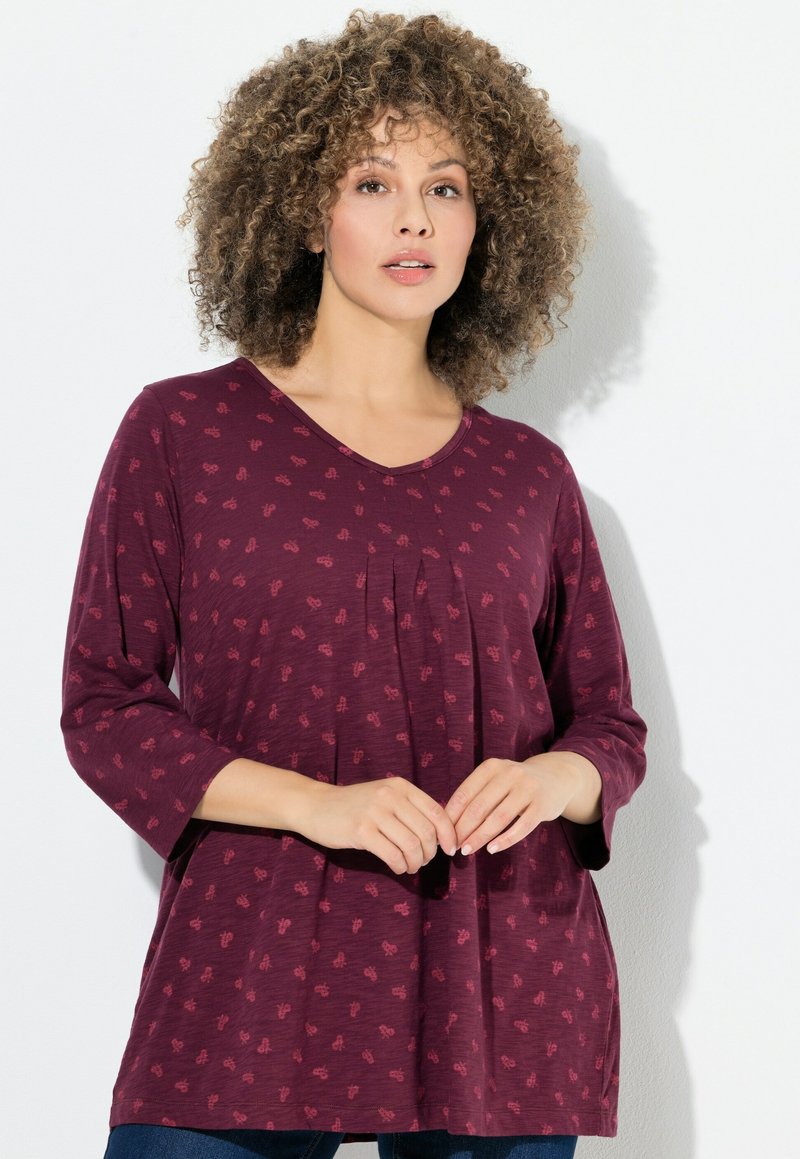 Top color bordeaux con maniche a tre quarti, scollo a V e pieghe frontali. Presenta un motivo sottile ed è realizzato in tessuto morbido.