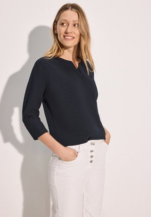 Haut texturé bleu marine avec des manches trois-quarts et un col discret, associé à un pantalon large blanc avec une fermeture à boutons.