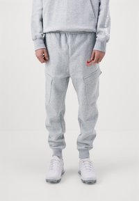 Šedé tepláky s uvolněným střihem, elastickým pasem, dvěma bočními kapsami a červeným logem Nike. V kombinaci s bílými sportovními botami.