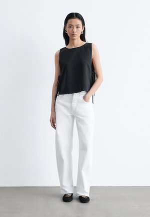 Kobieta stojąca, ubrana w czarny, bezrękawnik z wiązaniami po bokach, białe, szerokie spodnie z wysokim stanem oraz czarne baleriny, na tle białego tła.