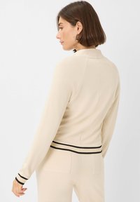 Vrouw met kort bruin haar, gekleed in een beige langemouwen top en bijpassende broek met zwarte accenten op de manchet en tailleband, die naar links kijkt.