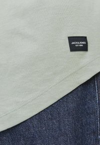 Világoszöld pamut póló, amelyen egy kis fekete címke található "JACK & JONES EST. 1990." felirattal. A lekerekített szegély sötétkék farmernadrággal találkozik, amely textúrált felületű.