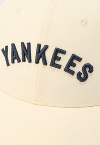 Gorra de béisbol beige con texto "YANKEES" bordado en negro en el panel frontal y detalles de costura visibles.