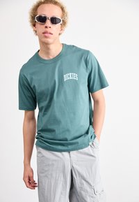 Teal színű pamut póló, rajta fehér "DICKIES" logóval, világosszürke cargo nadrággal párosítva. A modellen fehér ovális napszemüveg van.