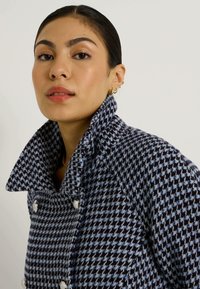 Manteau croisé en motifs pied-de-poule bleu marine et bleu clair, avec un col haut et des boutons argentés. Tissu doux et texturé.