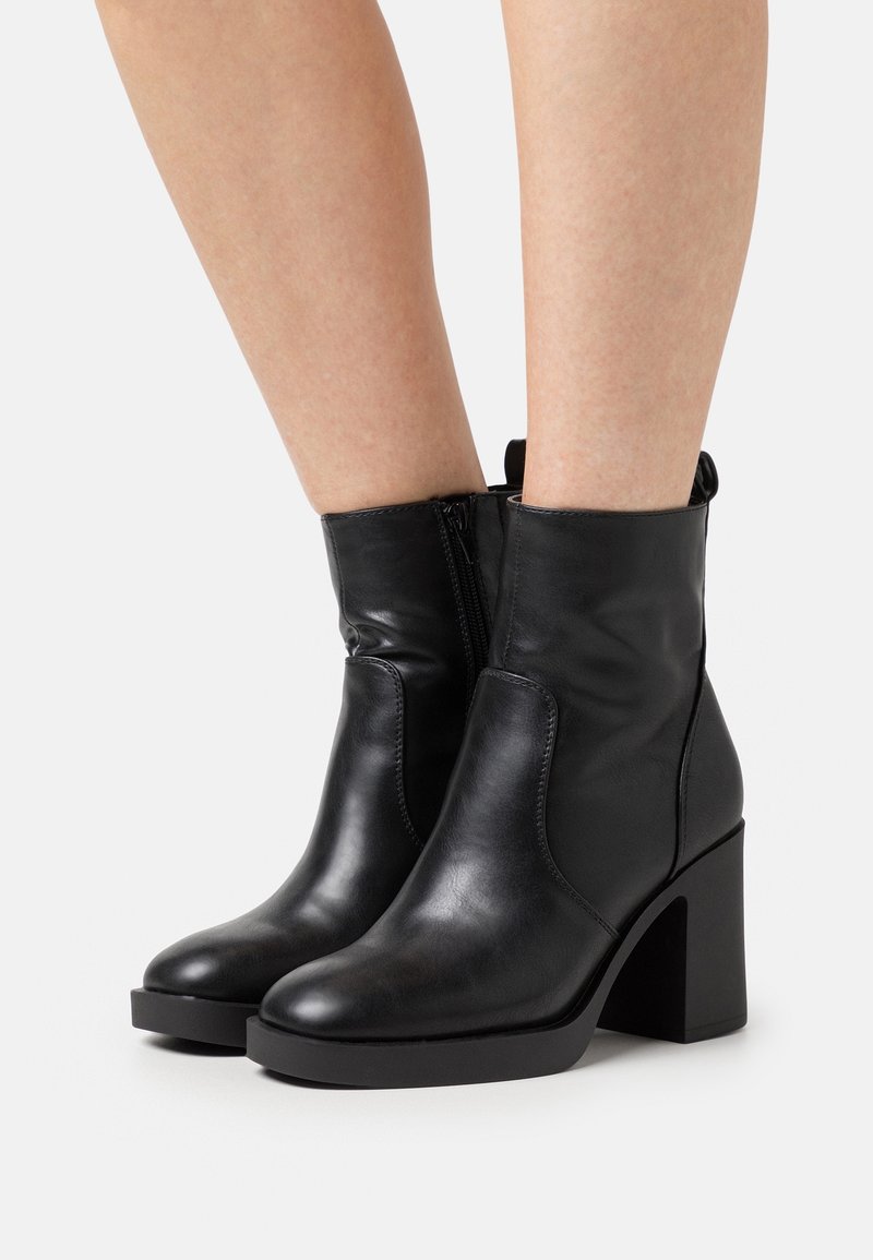 Bottines en cuir noir avec talons carrés portées sur des jambes nues, dotées de fermetures éclair latérales et d'orteils arrondis.