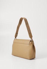 Beżowa torebka o fakturowanej powierzchni, z usztywnionym kształtem i paskiem z wyszywanym logo. Zdobiona srebrzystymi elementami metalowymi.