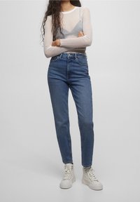 Höga blå jeans med avsmalnande ben, gjorda av denim. Kombinerat med en genomskinlig, texturerad långärmad topp och vita högstövlar.