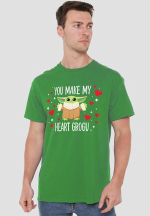 Young man in green t-shirt featuring Grogu character surrounded by red hearts and text saying "You make my heart Grogu."