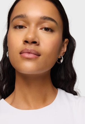 Jeune femme au teint moyen, cheveux ondulés foncés, portant des boucles d'oreilles créoles en argent et un haut blanc, expression neutre, fond clair.