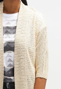 Jasnobeżowy sweter kardigan z krótkim rękawem i otwartym przodem, charakteryzujący się prążkowaną fakturą oraz luźnym, przewiewnym krojem.