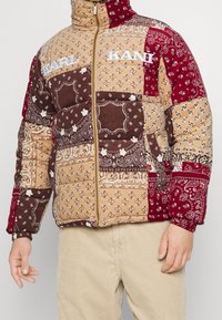 Gepolsterte Jacke mit Patchwork-Design in Hellbraun, Burgunderrot und Dunkelbraun mit Bandana-Mustern, hohem Kragen und Reißverschluss.