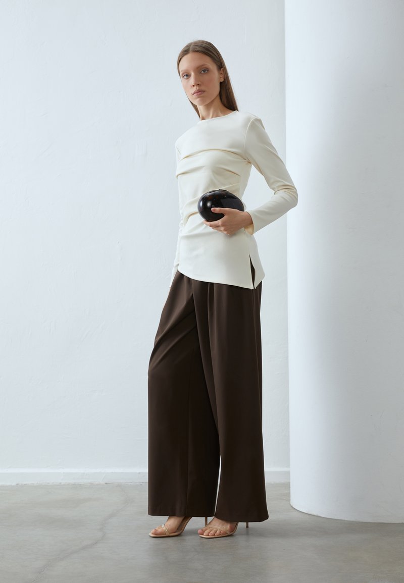 Biała bluzka z długim rękawem i marszczeniem po bokach, w połączeniu z szerokimi brązowymi spodniami. Modelka trzyma błyszczącą czarną torebkę, na tle minimalistycznym.