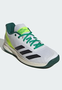 Scarpa da ginnastica bianca e verde con strisce nere, lacci verdi e branding giallo neon sul tallone e sulla linguetta, angolata per mostrare il lato esterno.