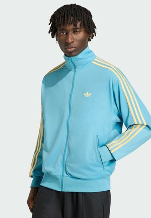 Mann mit Dreadlocks, der eine hellblaue Trainingsjacke mit Reißverschluss, gelben Streifen und Logo trägt, posiert mit einer Hand in der Tasche.