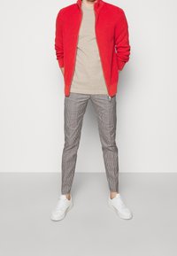 Červený, žebrovaný svetr na zip přes béžové tričko, spárovaný s kostkovanými zúženými kalhotami a bílými teniskami na neutrálním pozadí.