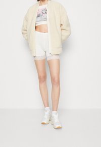 Veste bomber duveteuse couleur crème, crop top blanc, short blanc avec bordure en dentelle, chaussettes blanches et baskets blanches sportives.