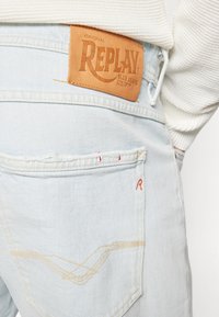 Jeans de mezclilla de color azul claro con un parche de cuero con el logo de REPLAY, con costuras decorativas en los bolsillos traseros y acentos de hilo rojo.