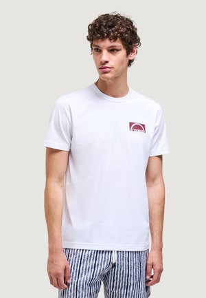 Giovane uomo con capelli ricci che indossa una maglietta bianca con un piccolo logo bordeaux sul petto e pantaloncini a righe con coulisse, che guarda di lato.