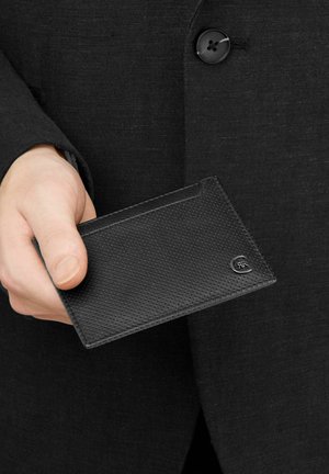 Ruka držící černý texturovaný kožený držák na karty s malým kovovým logem, oblečená v černém obleku s viditelným knoflíkem.