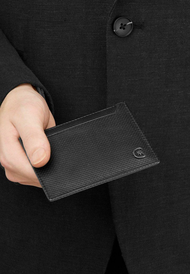 Mano sosteniendo un tarjetero de cuero negro texturizado con un pequeño logo metálico, vestido con una chaqueta de traje negra con botón visible.