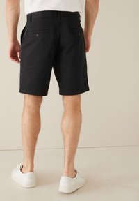 Schwarze Shorts mit glatter Textur, zwei Gesäßtaschen mit Knopfverschlüssen und geradem Schnitt. Getragen mit weißen Turnschuhen.