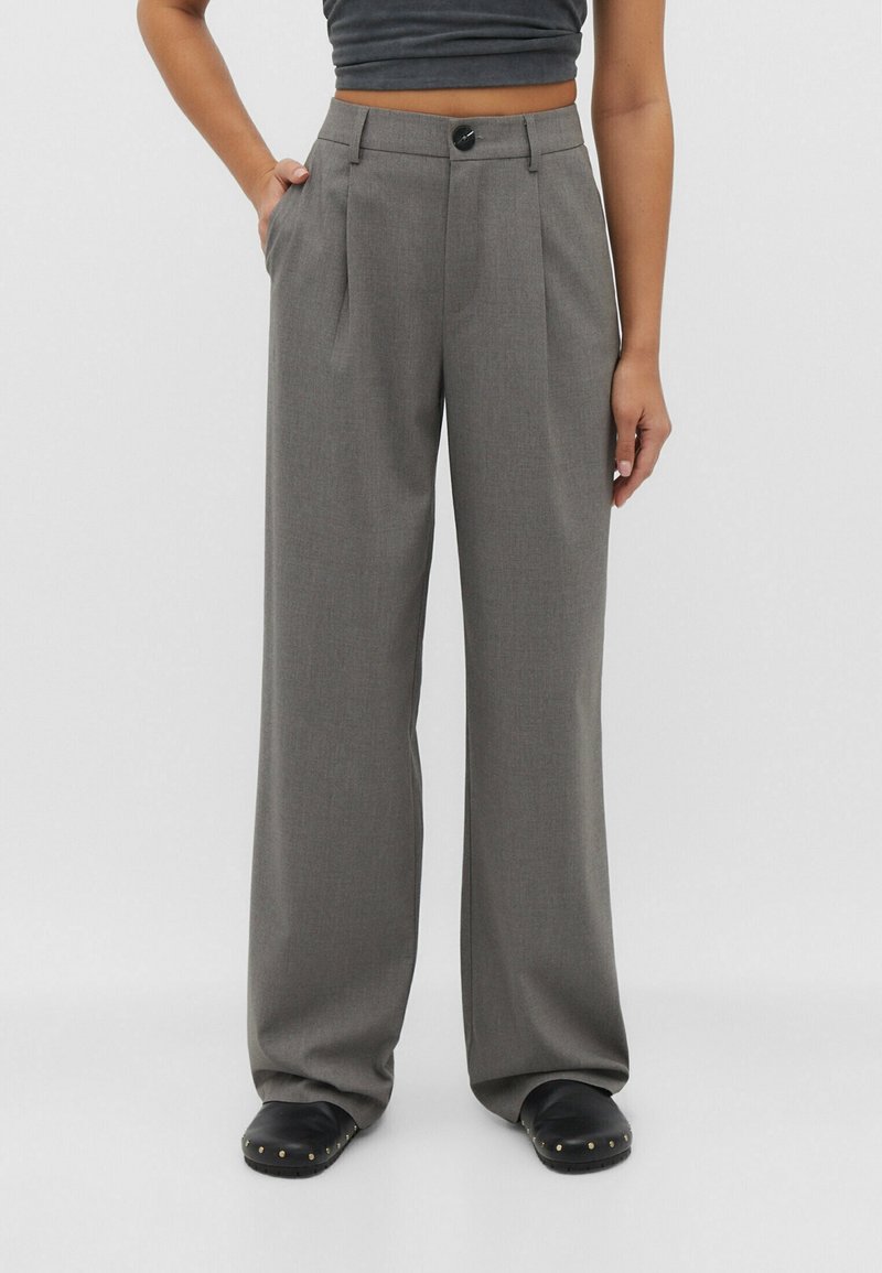 Pantalon gris sur mesure à jambes larges avec plis devant, bouton à la taille et poches latérales. Assorti à des sabots noirs ornés d'accents dorés.