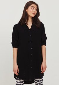 Schwarzes Hemdkleid mit weitem Kragen, kurzen Puffärmeln und einem längeren Rückensaum. Hergestellt aus einem glatten Stoff mit einer leicht durchscheinenden Textur.