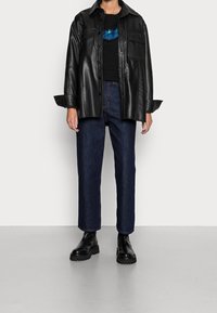Persona vestida con una chaqueta de cuero negra, camiseta negra con diseño azul, jeans oscuro azul y botas negras gruesas frente a un fondo sencillo.